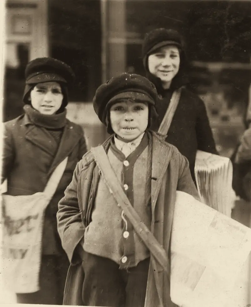 Three newsboys with canvas bags full of papers stand on a city street.