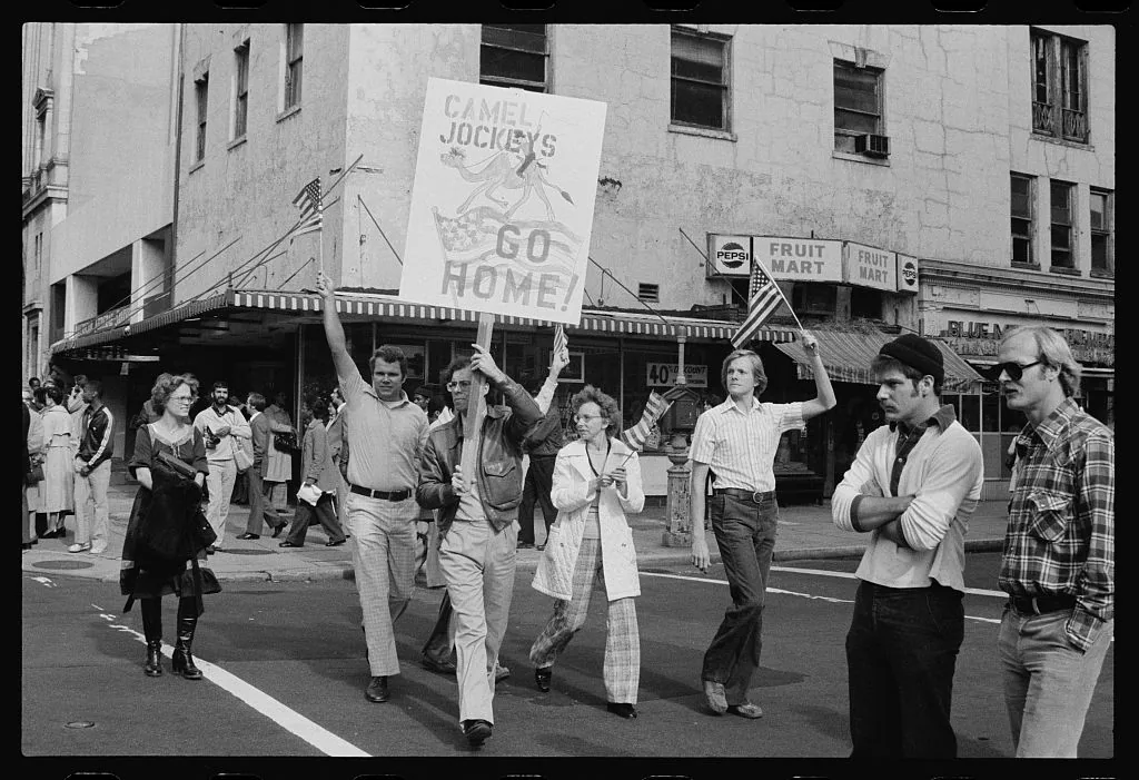 Demonstrators march down a city street in 1979 carrying an American flag and a hand lettered sign that uses an ethnic slur and tells Iranians to go home
