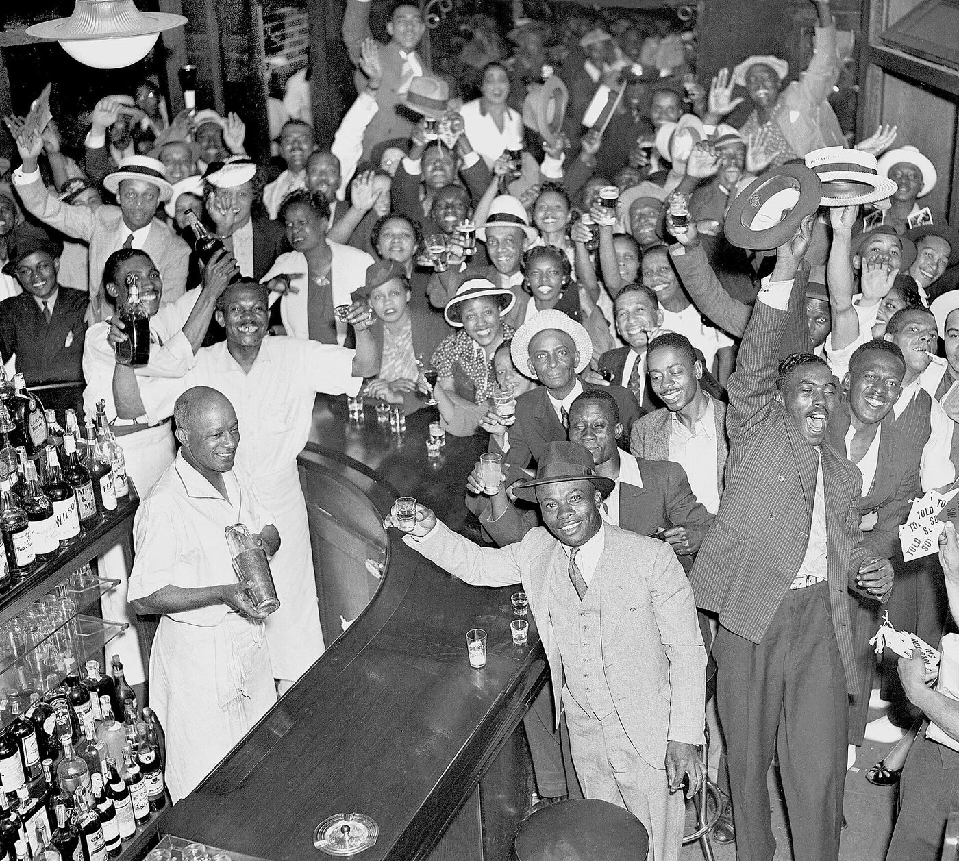 A packed bar in Harlem full of Black men and women cheering with raised arms and hats.