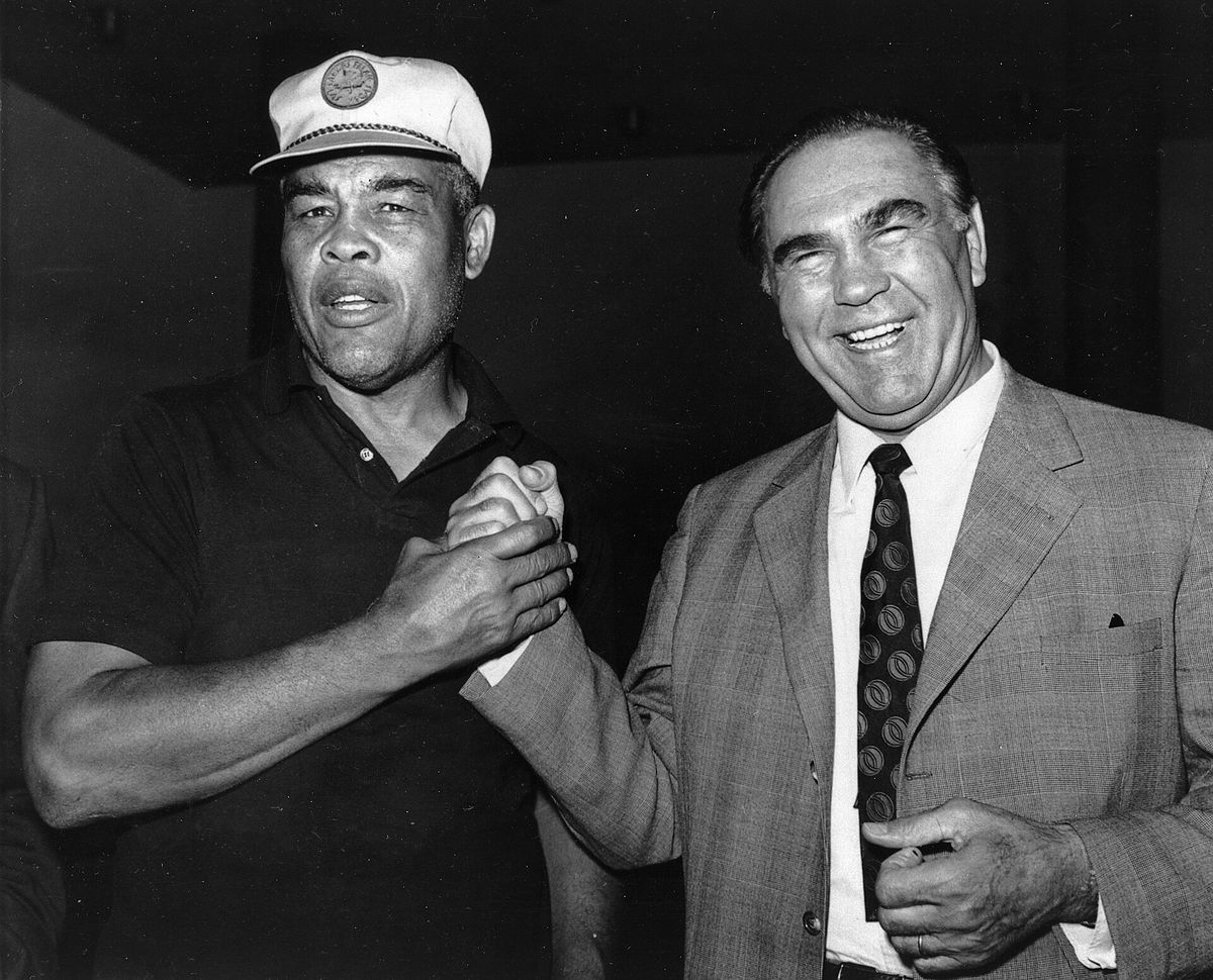 Joe Louis in a cap and Max Schmeling in a suit shaking hands and smiling.