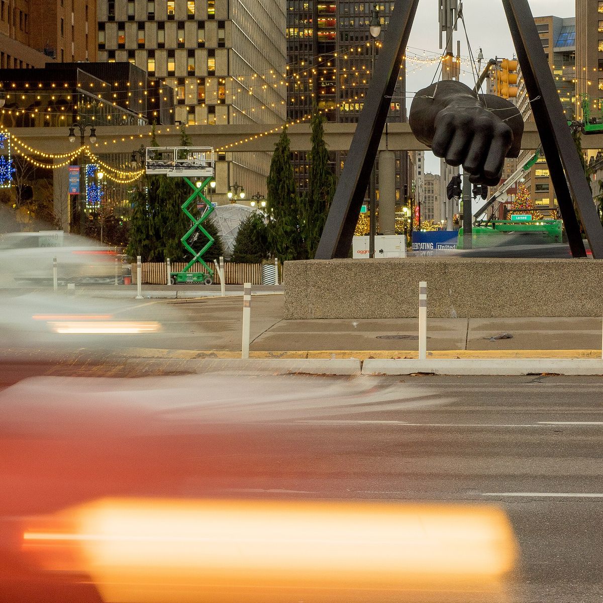 A giant bronze arm and fist hanging from a pyramid frame over a Detroit intersection at night.
