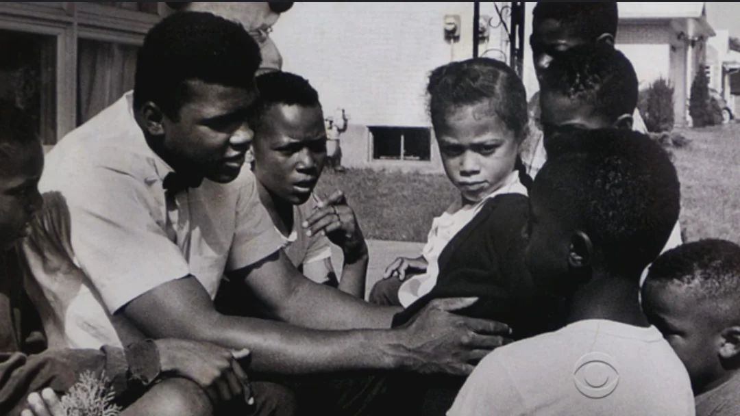 Muhammad Ali crouching and talking with a group of Black children outdoors.