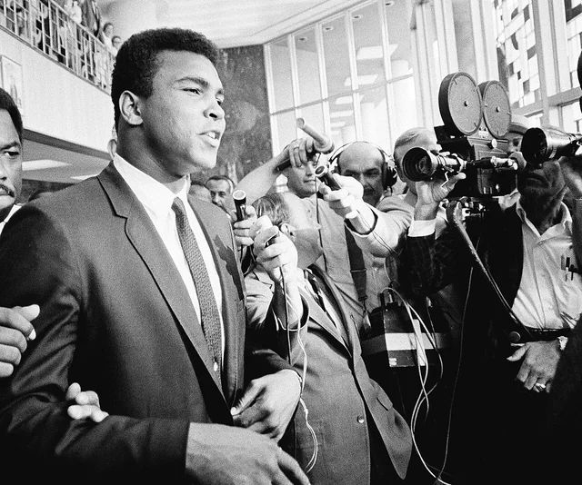 Muhammad Ali in a suit speaking in front of a bank of newsreel cameras and microphones.