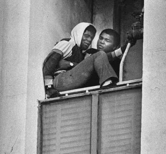 Muhammad Ali leaning out of a high window with his arm around a young man seated on a ledge.