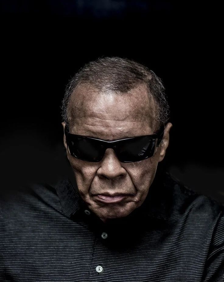 Muhammad Ali in later life, wearing dark sunglasses against a black background.