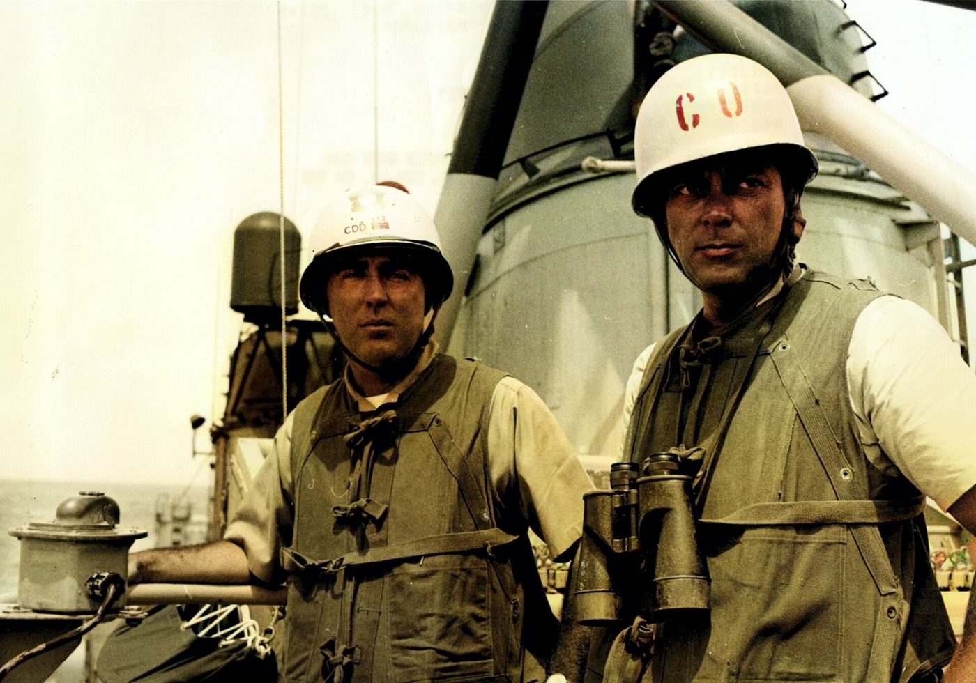 Two Navy officers in helmets and life vests on the deck of a destroyer. The man on the right wears a helmet marked CO.