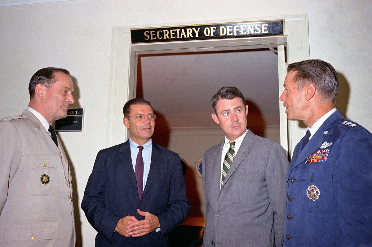 Four men in suits and military uniforms talking in a Pentagon doorway under a sign reading Secretary of Defense.