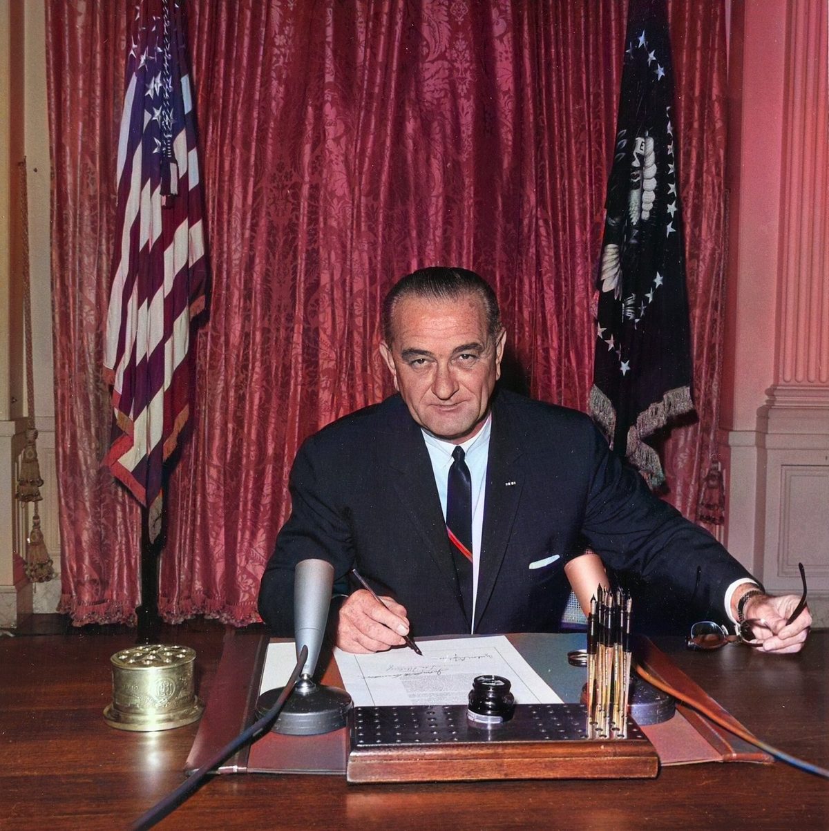 President Johnson seated at a desk in the East Room signing a document, flags and red drapes behind him.