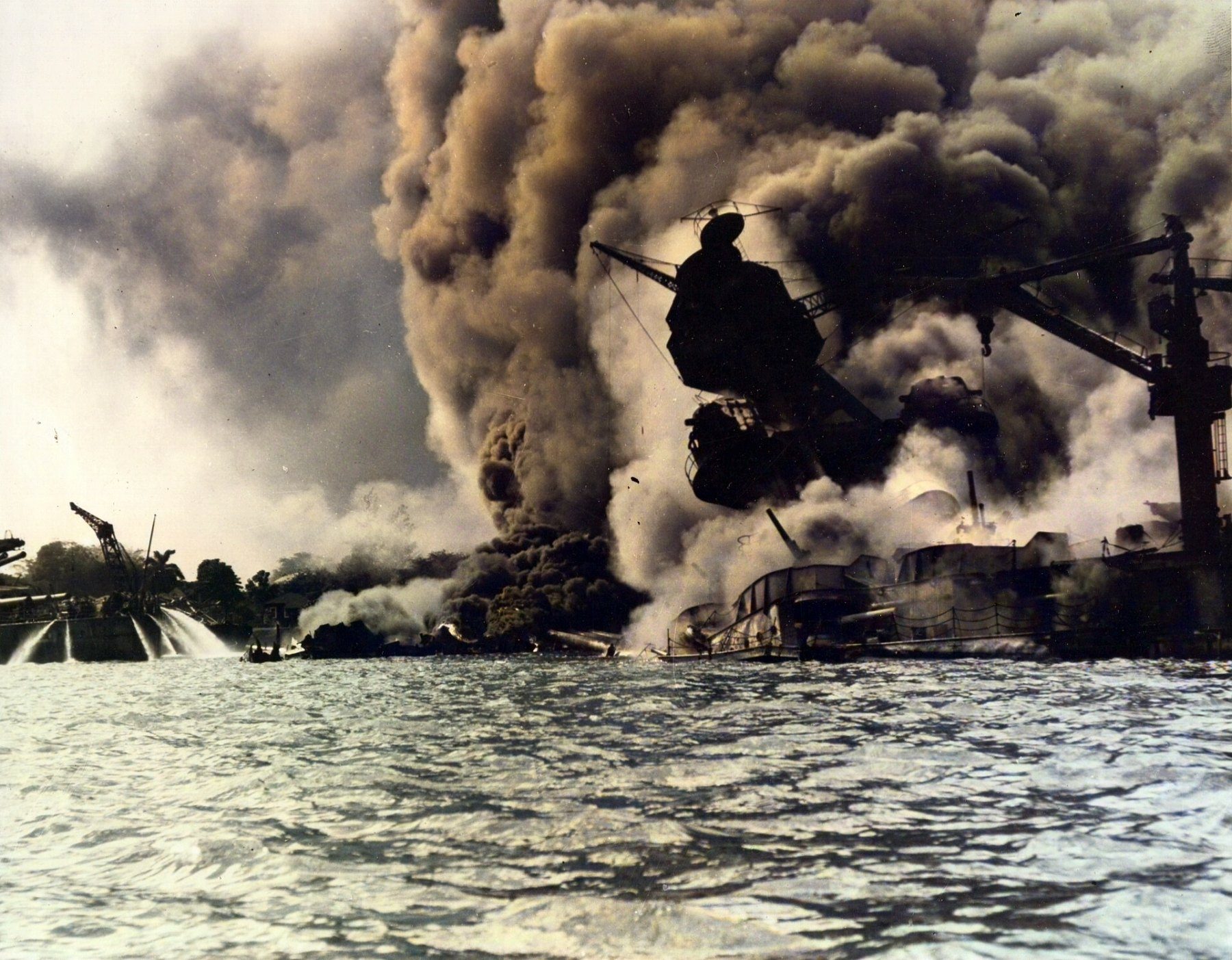 The battleship USS Arizona burning and sinking in heavy black smoke, with fire hoses spraying water in the foreground.