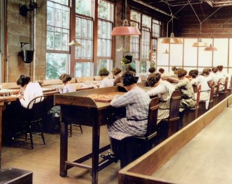 Rows of women sit at long wooden work tables in a bright factory room with tall windows.