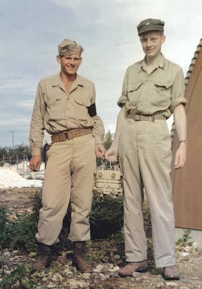 Two men in khaki uniforms stand outdoors on either side of a small metal carrying case.