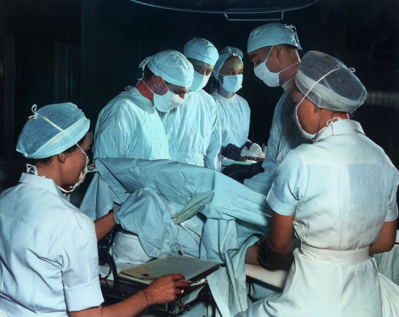 Masked surgeons and nurses in caps and gowns crowd around a patient on an operating table.