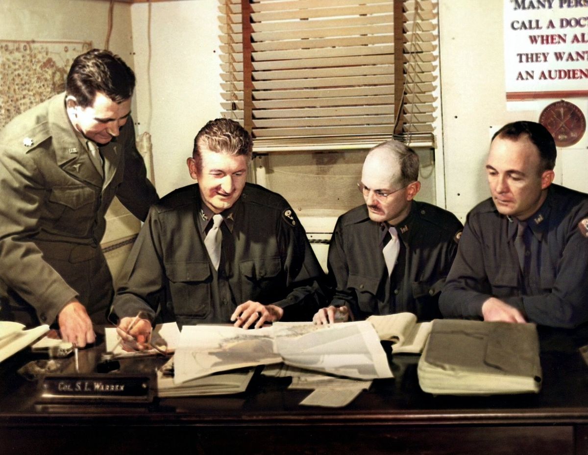 Four men in Army uniforms lean over papers spread across a desk with a nameplate reading Col. S. L. Warren.