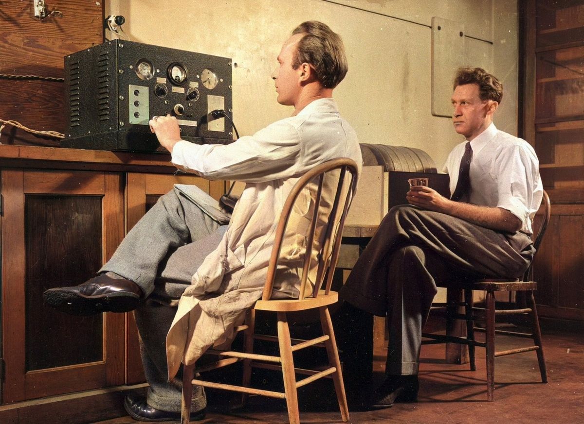 A man in a lab coat sits at a control panel with dials while a second man in a white shirt and tie sits beside him holding a small container.