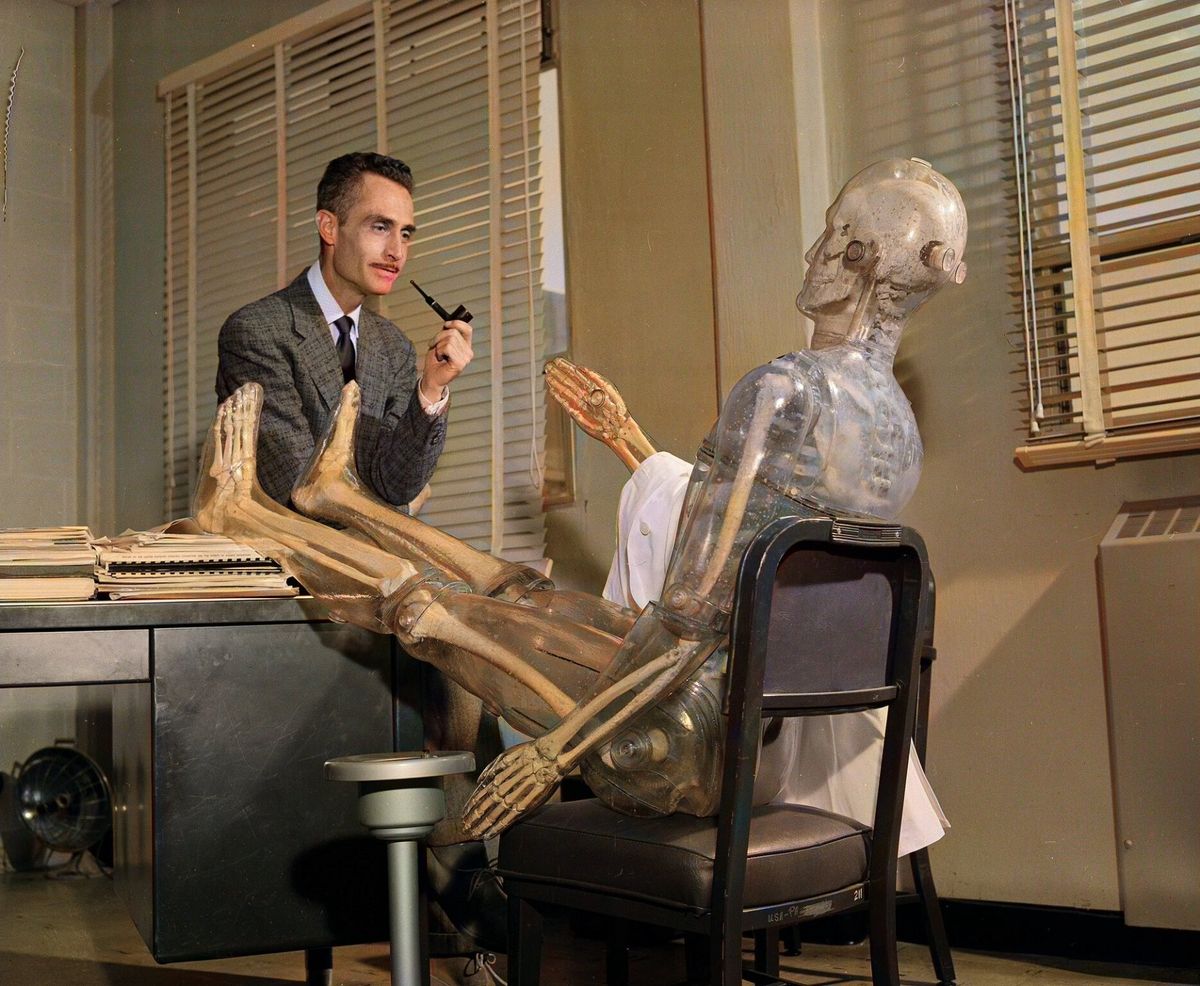 A man in a suit smoking a pipe sits at a desk beside a clear plastic model of a human body seated in a chair.