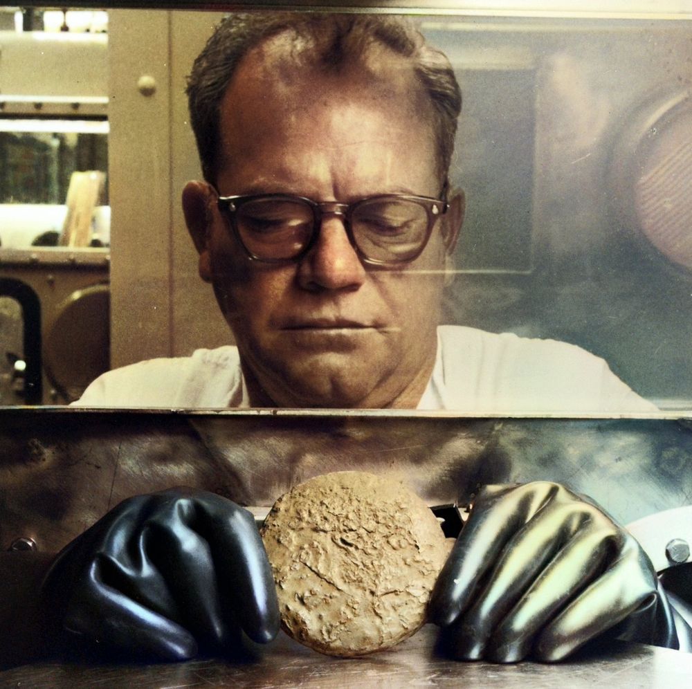 A man in glasses looks through a glovebox window at a round, rough metal disk held between two thick black rubber gloves.