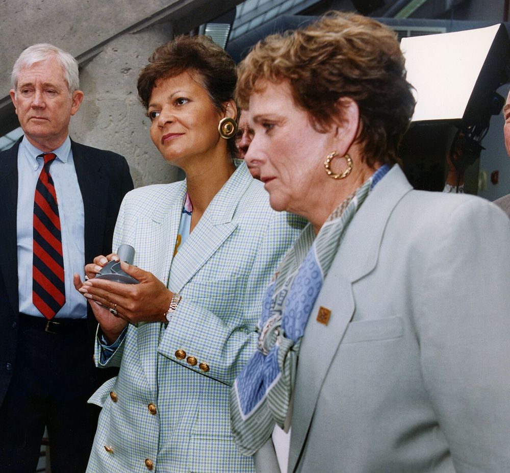 Two women in light suits and a man in a dark suit look intently at a small object one of the women holds.