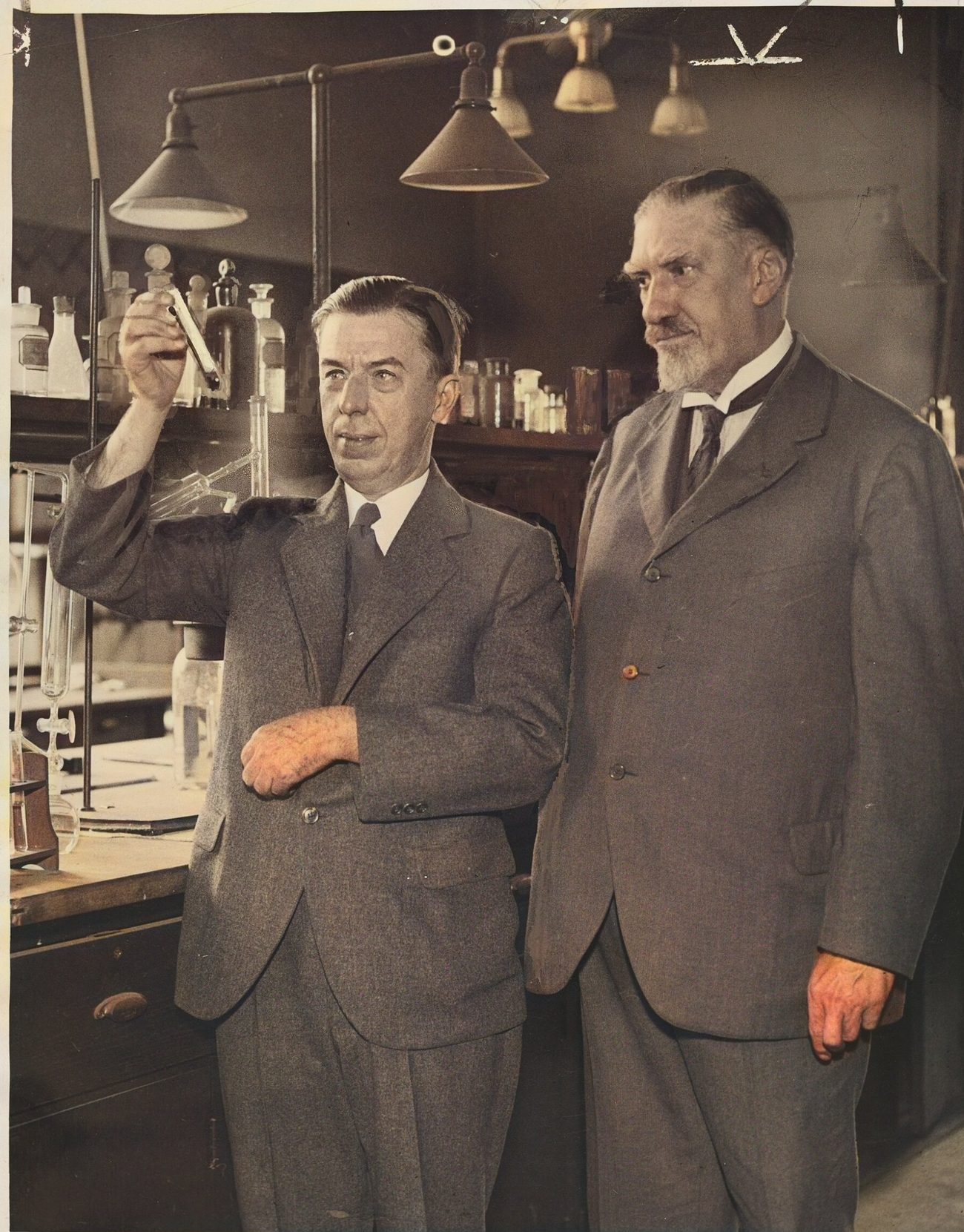 Two men in suits stand in a laboratory crowded with glassware and hanging lamps. The younger man, left, holds a flask up to the light.
