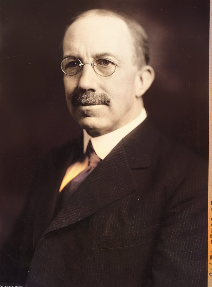 Formal portrait of a man with round wire glasses and a trimmed mustache.
