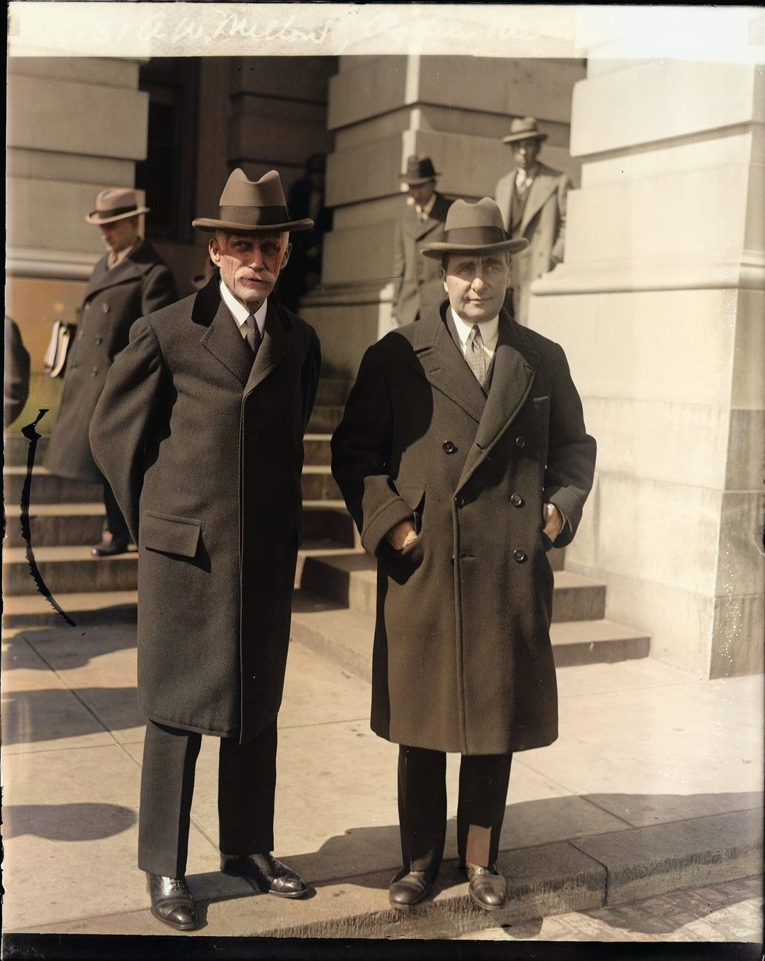 Two men in dark overcoats and hats stand on the steps outside a stone government building.