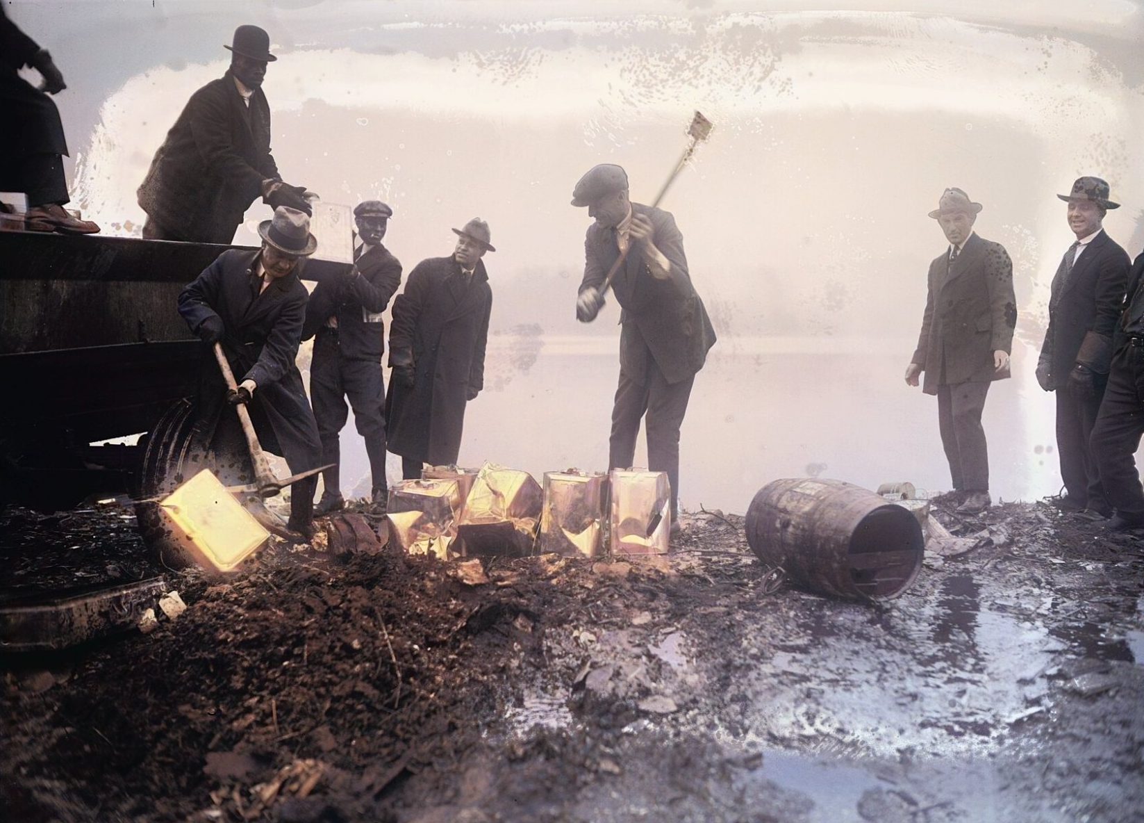Men in hats and overcoats, dark against a hazy sky, swing axes at containers of seized liquor on muddy ground.