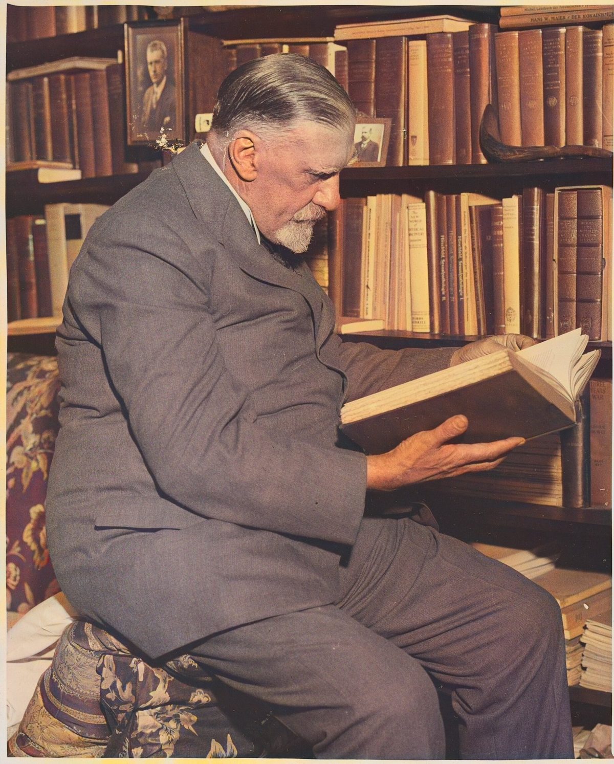 An older man with a white beard sits reading a large book in front of tall shelves of books.
