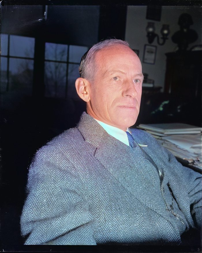 A lean man in a tweed suit sits at a desk and looks toward the camera.