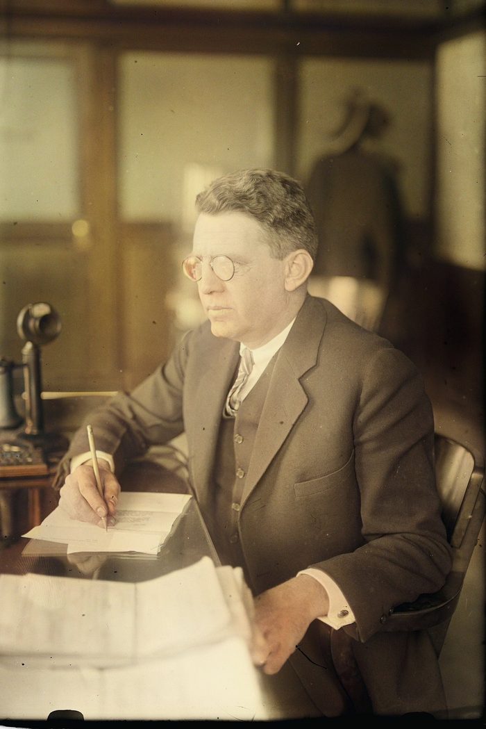 A man with round glasses writes at a desk beside an upright telephone.
