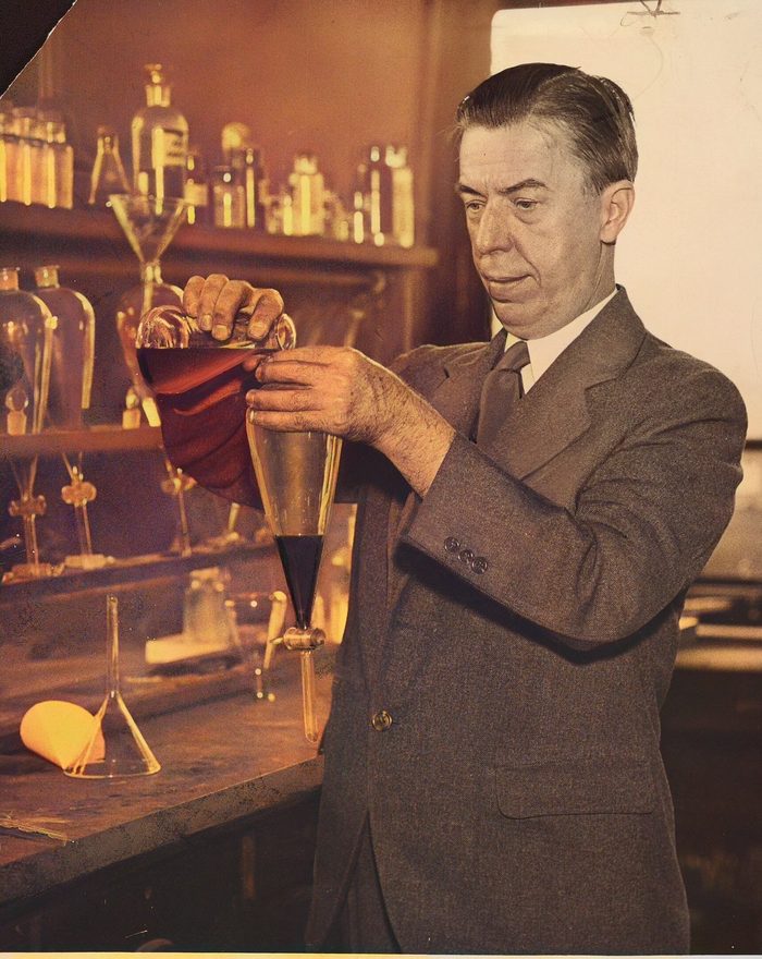 A man in a suit pours liquid through a funnel into a flask at a laboratory bench lined with bottles.