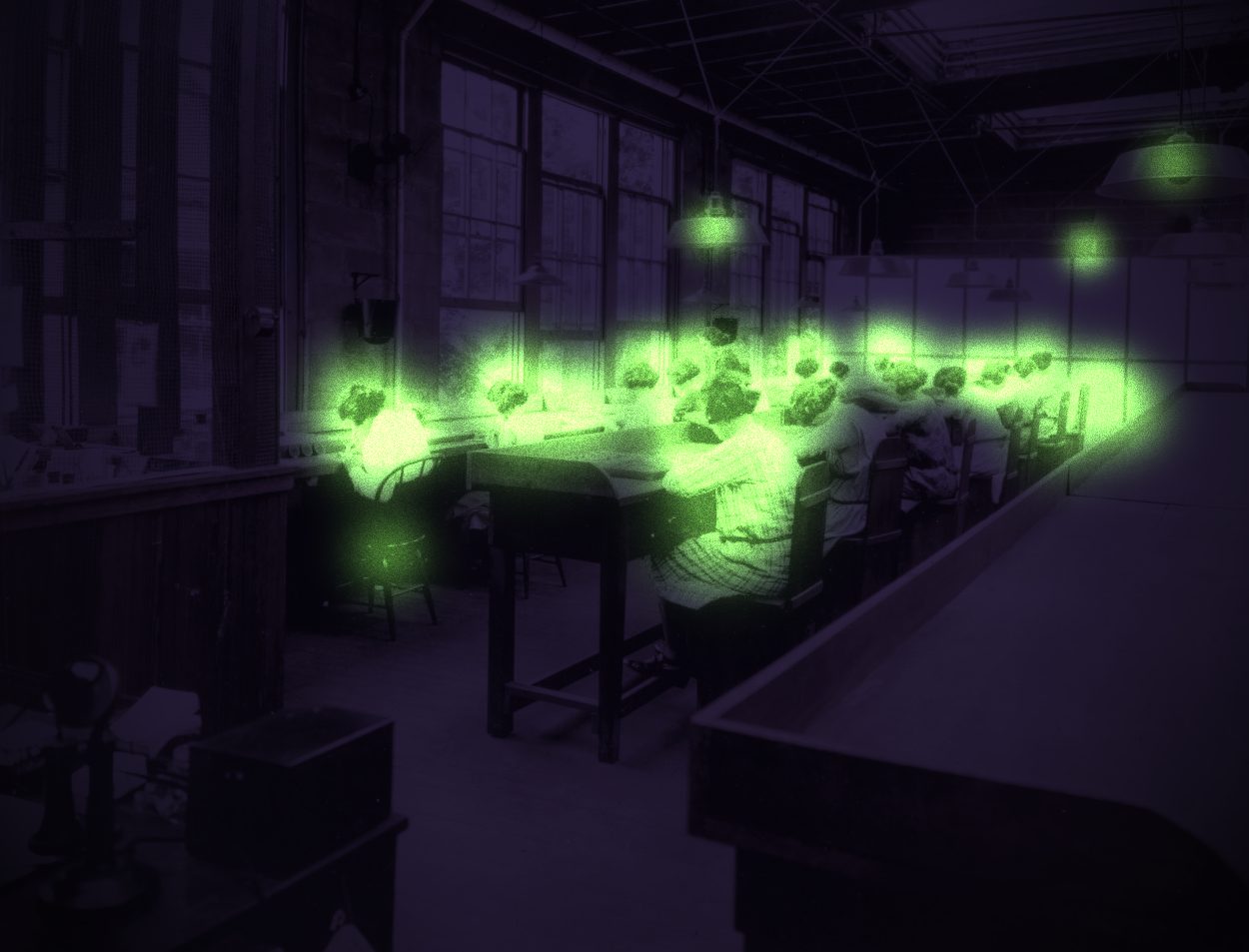 A row of young women at work tables in a darkened factory studio, their hair, hands and trays glowing green