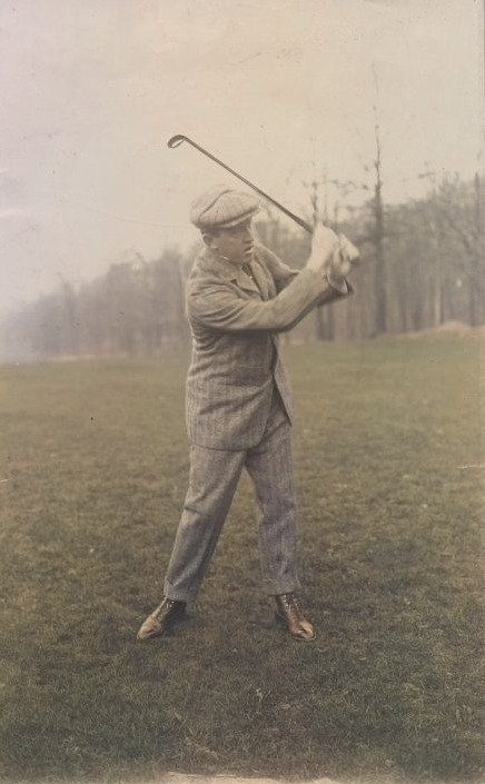 A man in a cap and tweed suit finishes a golf swing on a grassy course.