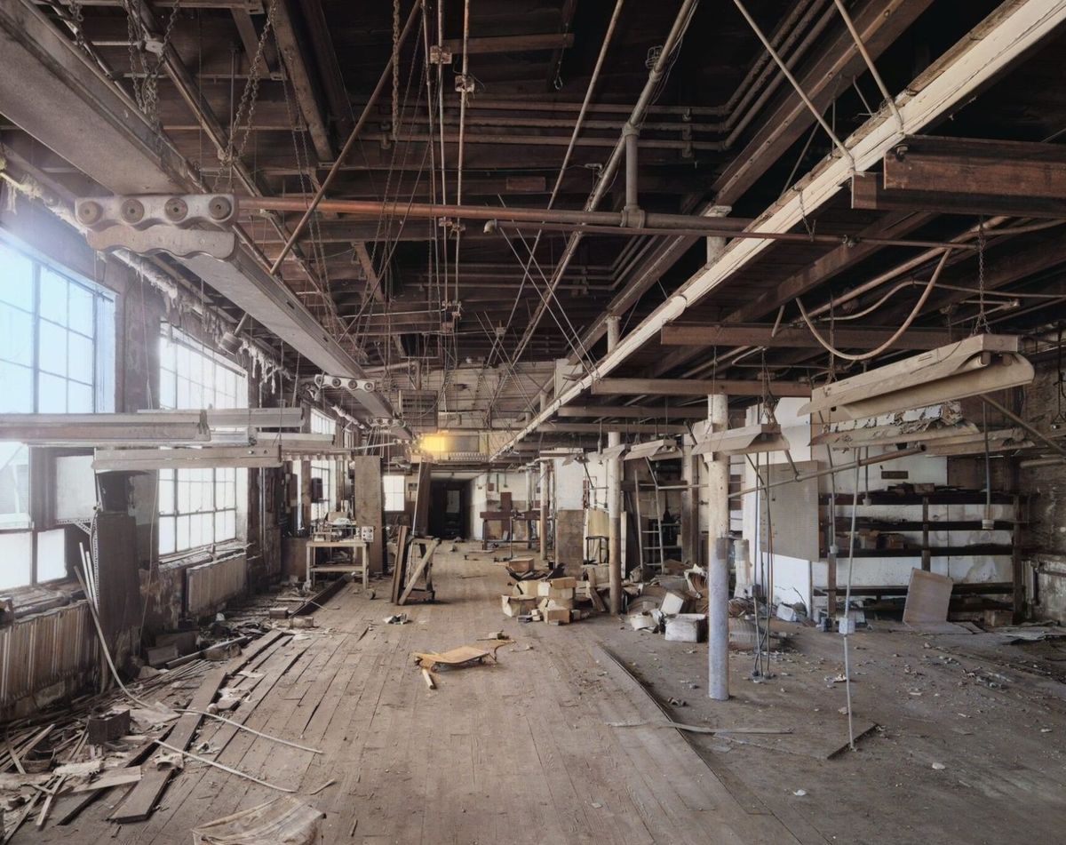 An empty second floor factory room with broken boards, dangling wires and tall dirty windows.