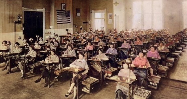 A large studio room packed with rows of young women at small desks with lamps, trays of dials beside them and an American flag on the back wall.