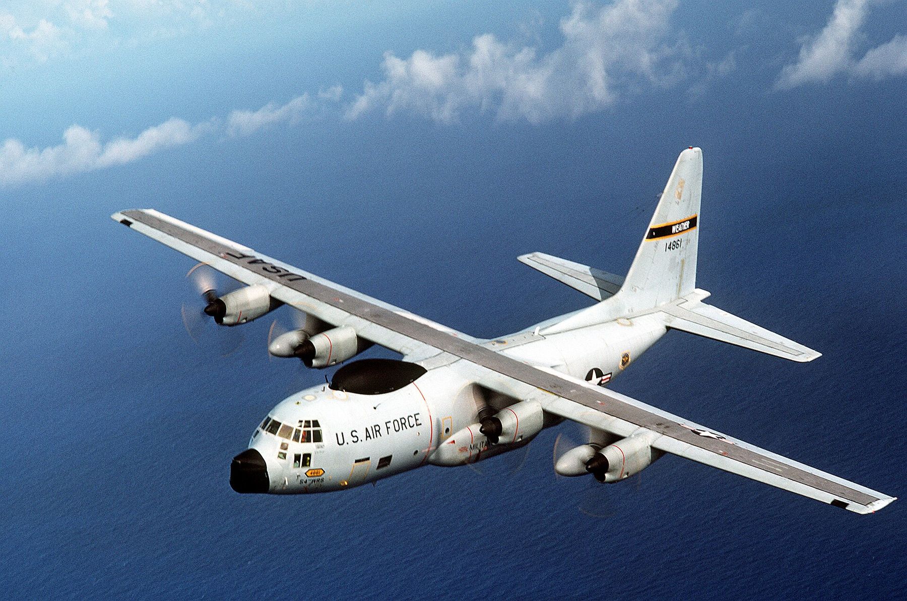 A white Air Force WC-130 weather reconnaissance plane flying over a blue ocean dotted with clouds