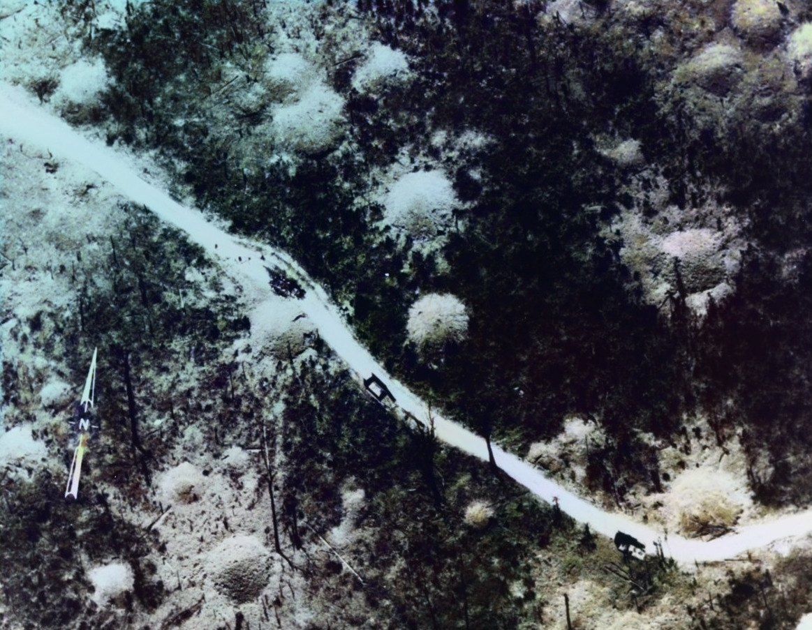 Aerial photograph of a pale dirt road cutting through dark jungle and bomb craters. White printed labels read unnumbered road segment and destroyed truck, and a circle marks a spot in the trees.