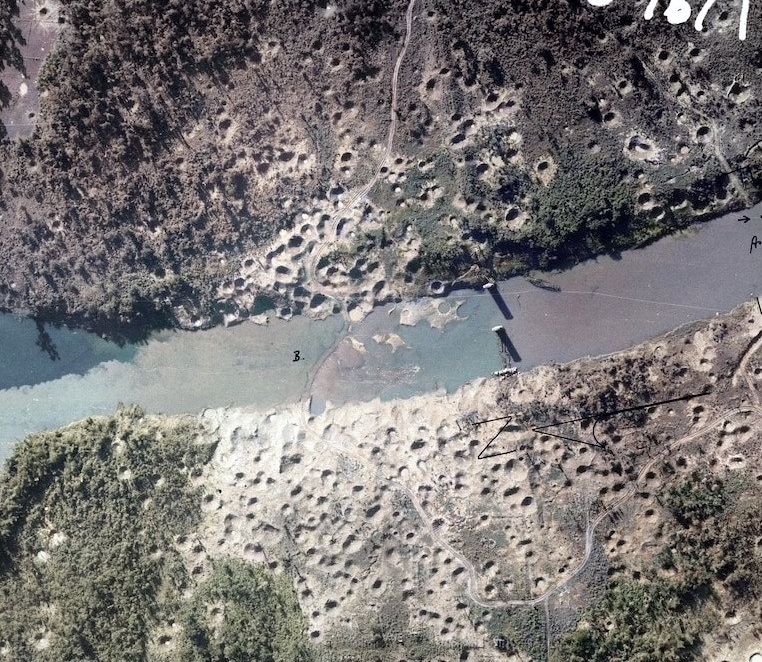 Aerial photograph of a river crossing surrounded by hundreds of round bomb craters on bare ground.