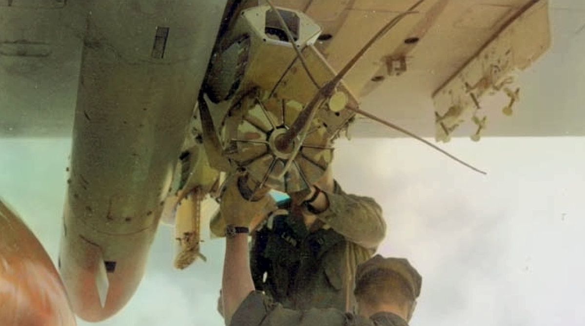 Looking up at the underside of a Navy patrol plane as a crewman lifts a long cylinder into a rack under the wing.