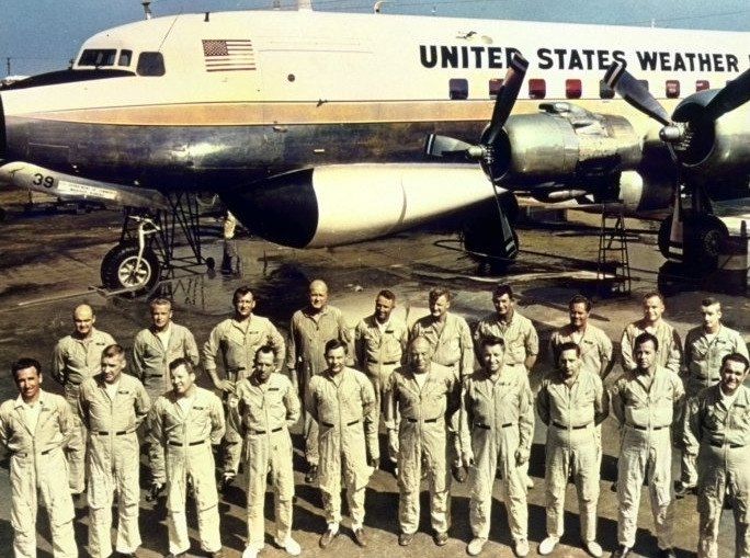 About twenty men in flight suits stand in rows in front of a large propeller airliner marked United States Weather Bureau.