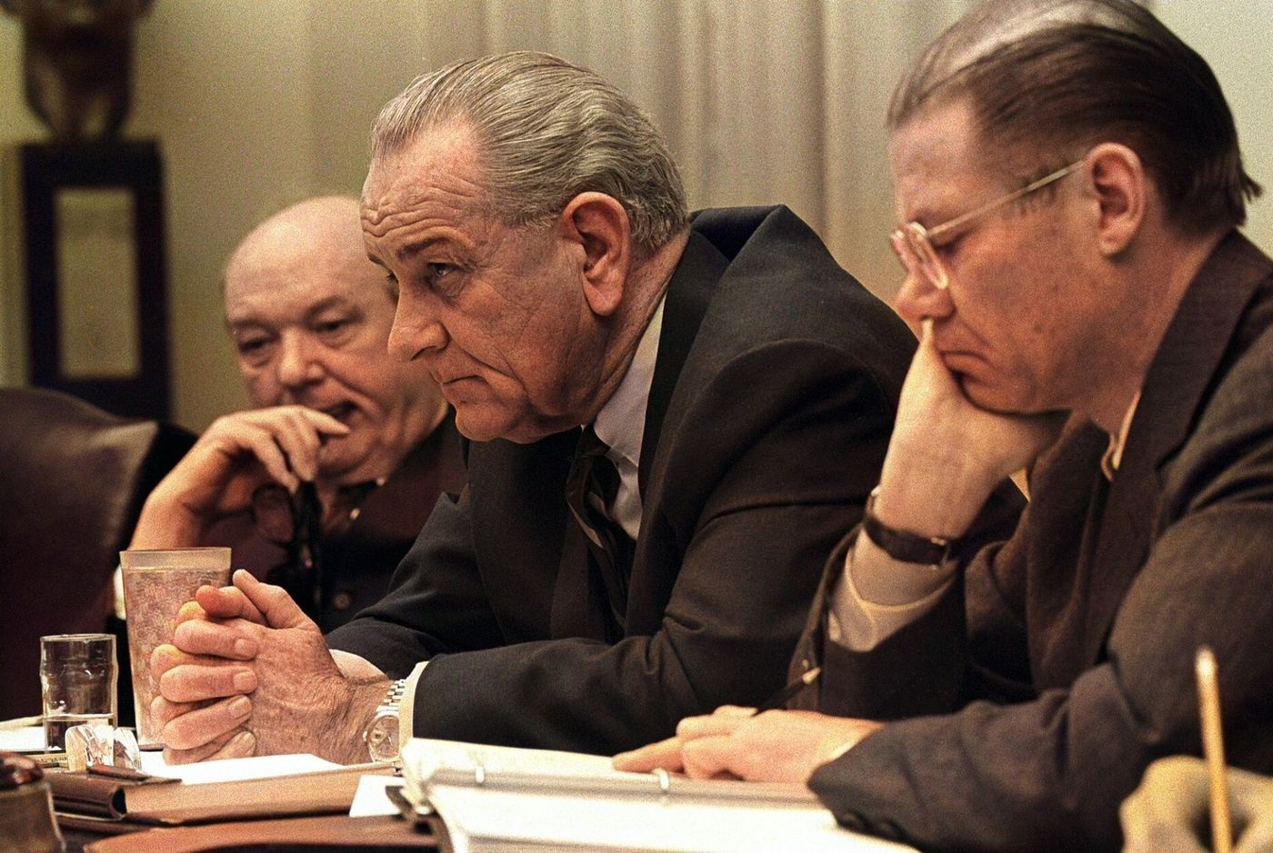 Three men in dark suits sit close together at a long table. The man in the middle leans forward with his hands clasped and looks off to the side.