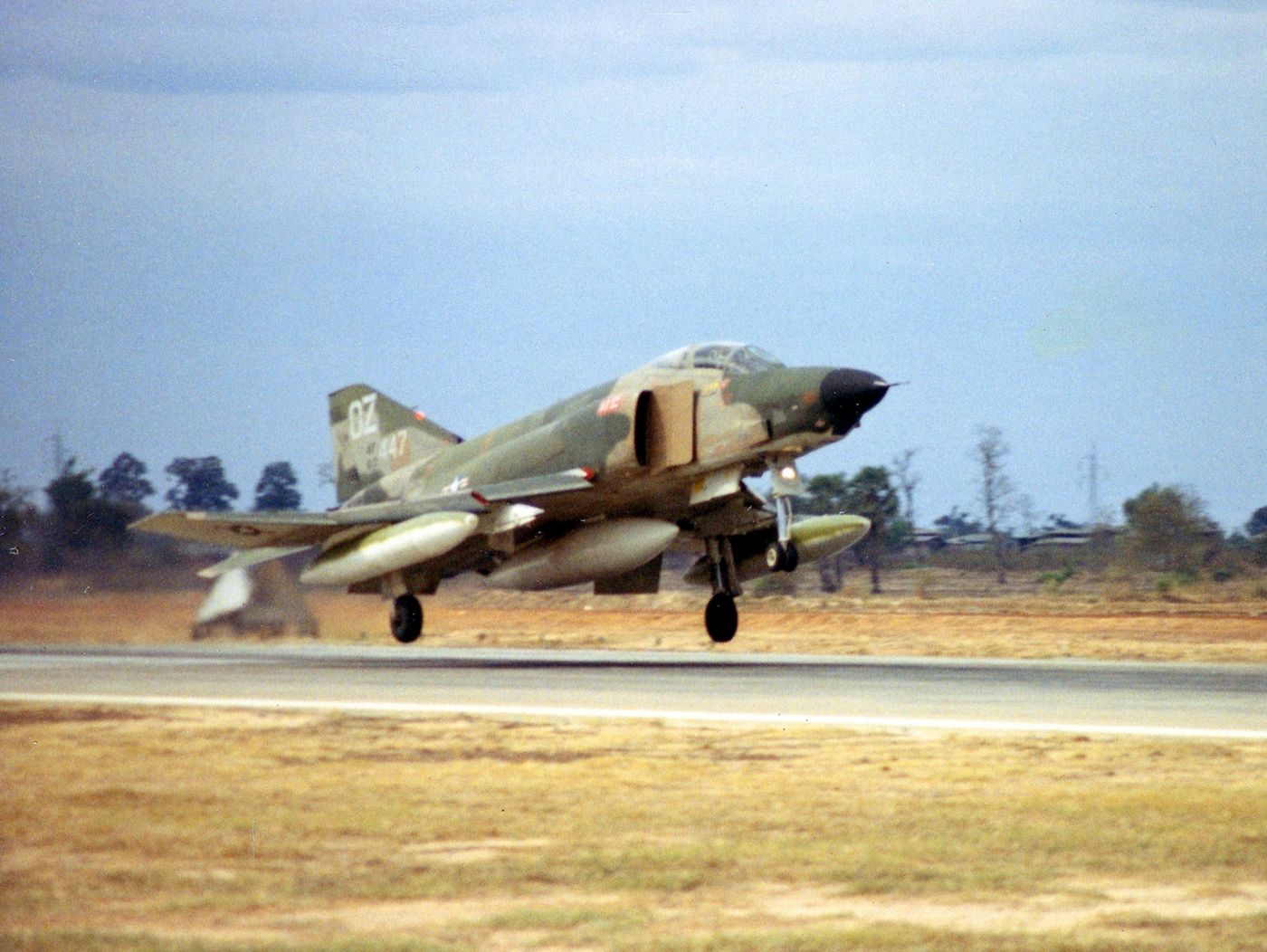 A camouflaged jet fighter with its landing gear down touches down on a runway past dry grass and palm trees.