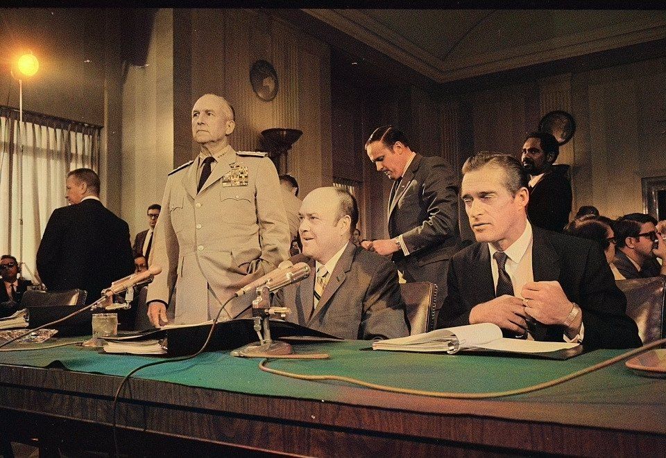 A man in a suit sits at a witness table covered with microphones in a paneled hearing room, with a senior officer in a light uniform standing behind him.