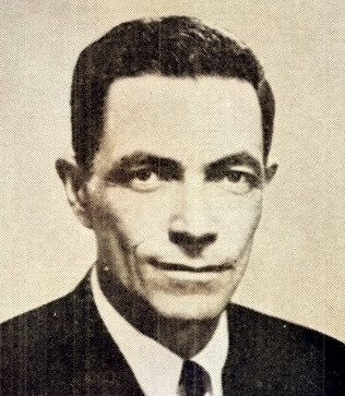 Portrait of a lean man with dark combed hair in a dark suit and tie.
