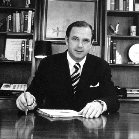 Alexander Butterfield in a suit writing at a desk in front of bookshelves.