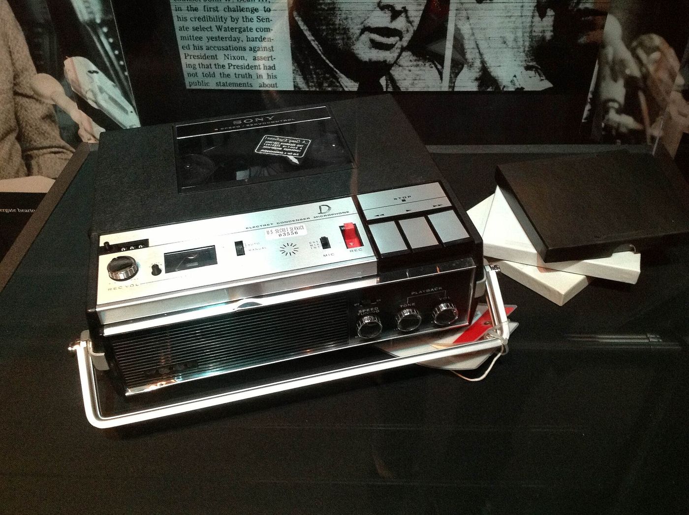 A silver reel to reel tape recorder with control buttons, displayed in a museum case.