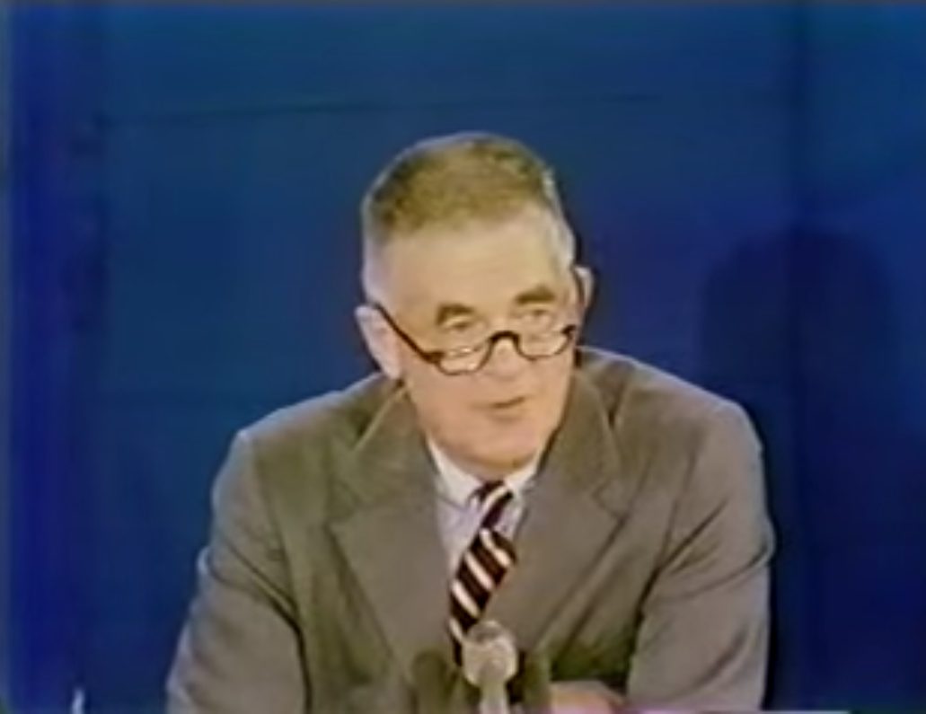 Archibald Cox in glasses and a bow tie speaking at a press conference, from a color television feed.