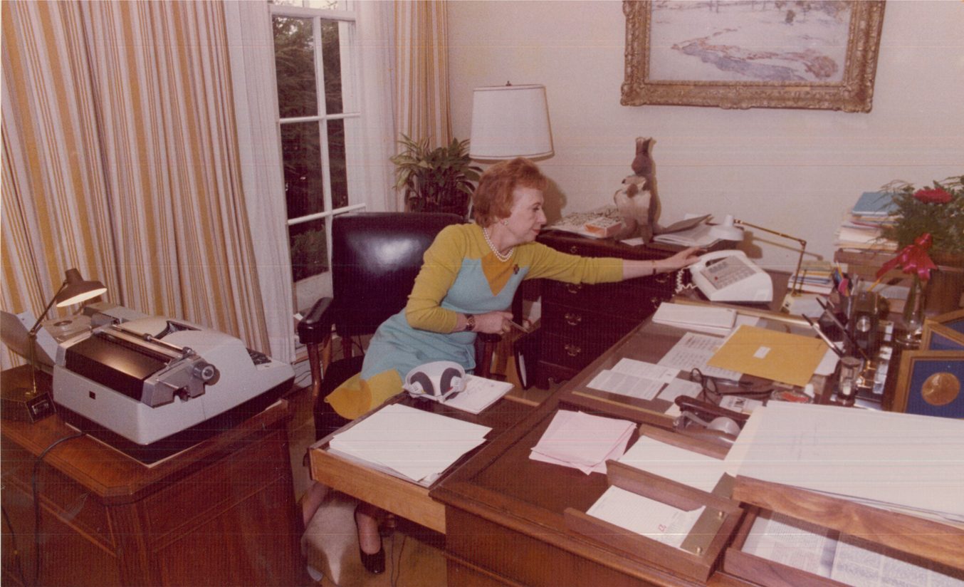 Rose Mary Woods at her office desk stretching her arm far to the left toward a telephone while her foot reaches toward a pedal under a tape recorder.