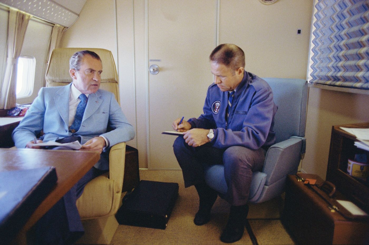 President Nixon and H. R. Haldeman seated facing each other in the cabin of Air Force One, Haldeman taking notes.