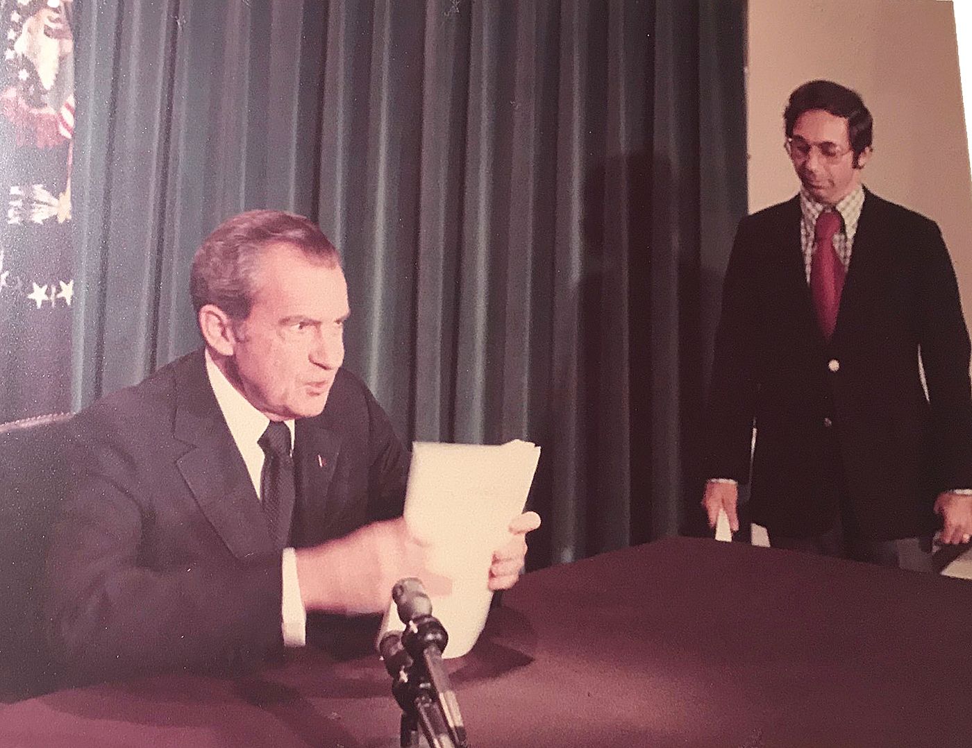 Nixon seated at a desk holding the pages of his speech, an aide standing behind him, before the television address.