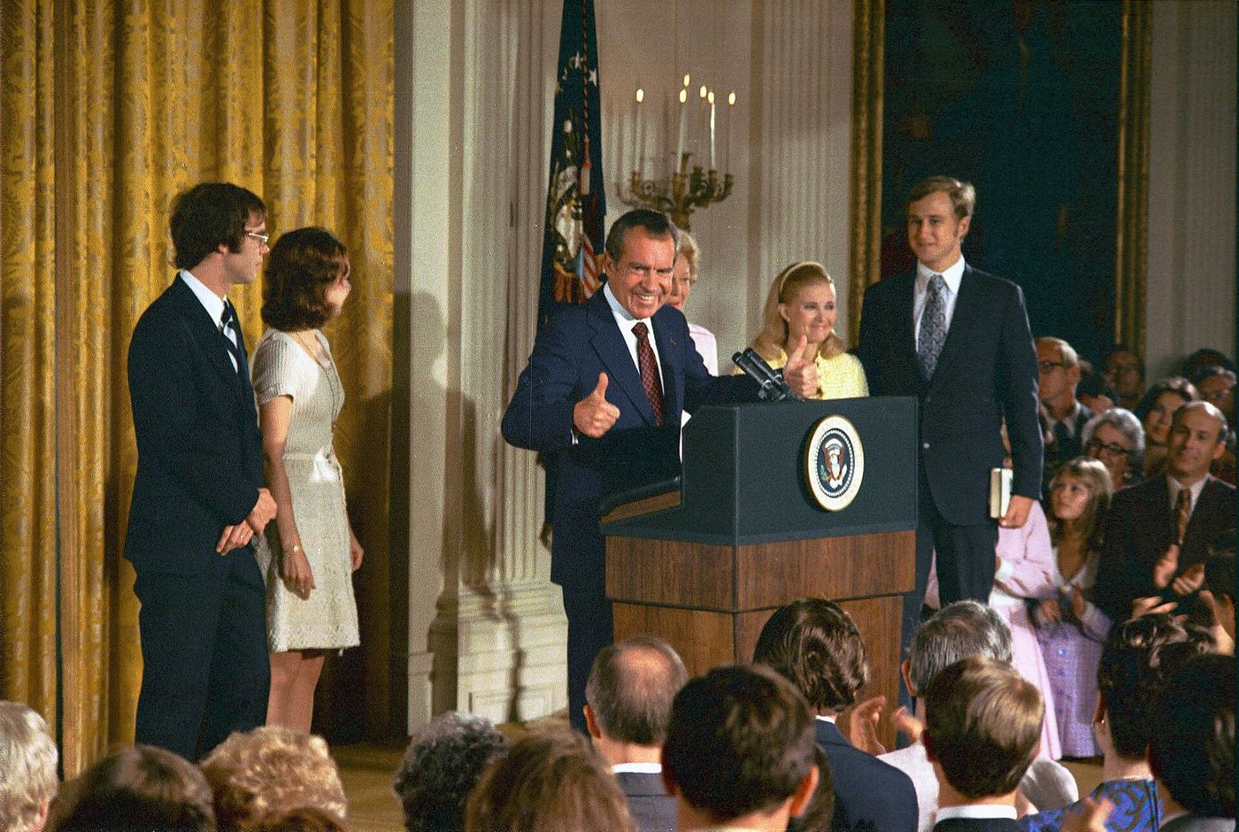 Nixon at a podium in the East Room with his family beside him and a crowded room of staff.