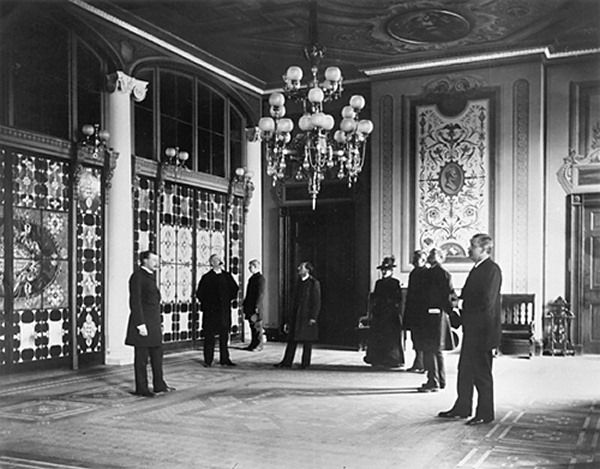 Black and white photograph of a grand hall with chandeliers and a tall screen of colored glass panels between columns